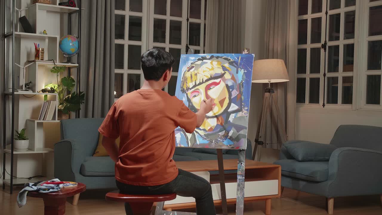 Hind View Of An Asian Artist Boy Sitting On Round Wooden Chair Without Backrest Holding Paintbrush Mixed Colour And Painting A Girl'S Face On The Canvas