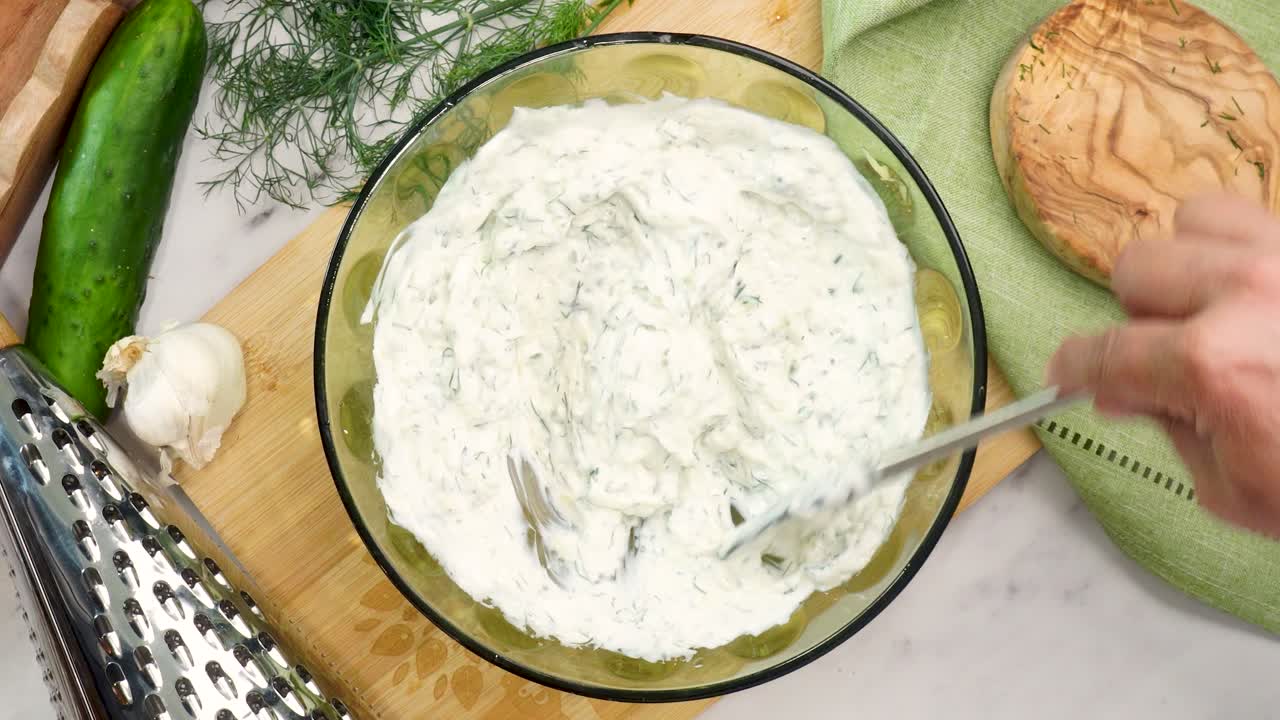 Making Tzatziki Greek or Mediterranean sauce or dip for a party. Dip or sauce in a green bowl on a wooden cutting board with dill, green napkin and a grater. Mixing the Tzatziki sauce for all foods.