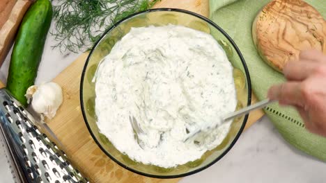 making tzatziki greek or mediterranean sauce or dip for a party. dip or sauce in a green bowl on a wooden cutting board with dill, green napkin and a grater. mixing the tzatziki sauce for all foods.