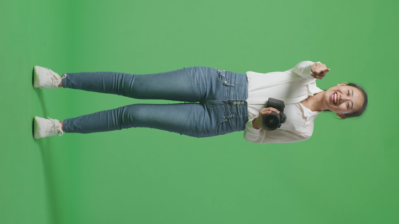 사진을 찍고 미소 짓고 그녀의 가을 만지는 카메라를 사용하는 아시아 사진작가의 몸 전체가 스튜디오에서 녹색 스크린 배경에 서있는 동안