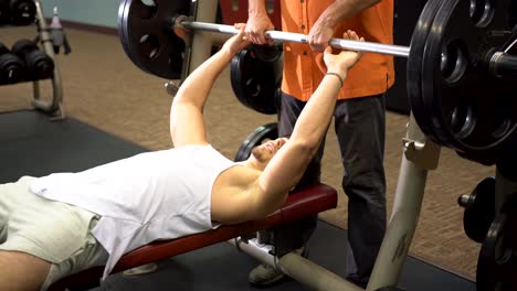 teen male bodybuilder lying down and pressing large barbell as a man in organge spots him