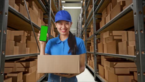 mensajera asiática en uniforme azul sonriendo y mostrando una pantalla verde del teléfono inteligente mientras entrega una caja en el almacén