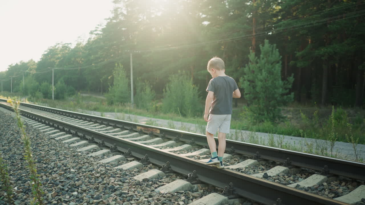 little boy walking carefully along rail track under bright sunlight with surrounding forest, gravel path, and overhead power lines in background