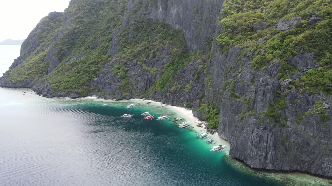 Breathtaking aerial view showcasing a secluded tropical cove nestled beneath towering limestone cliffs in El Nido, Palawan. Traditional Filipino bangkas boats anchored along the narrow shoreline