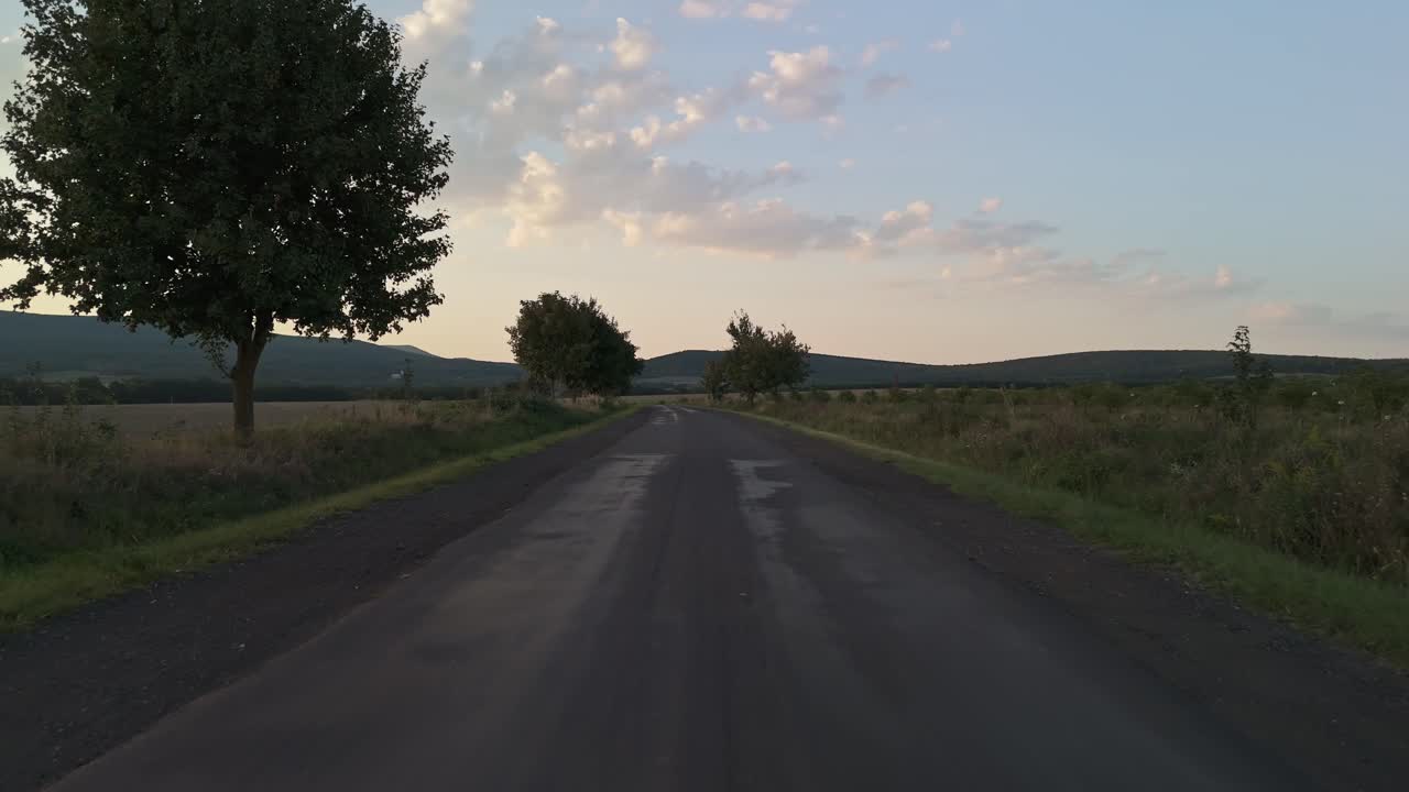 Ripresa a volo radente su una strada rurale dissestata che porta a Vilyvitány, Ungheria, regione di Zemplén al tramonto, delimitata da alberi e campi, con un segnale di pericolo attraversamento cervi che aggiunge contesto alla tranquilla campagna