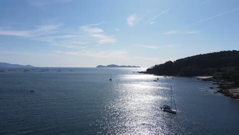 drone footage of ria de vigo, galicia. sunlight glistening on the calm sea, sailboats gently floating near the rugged coastline, with forested hills and distant islands under a clear blue sky.