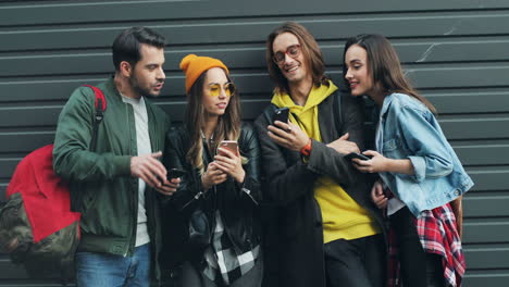 Group-of-caucasian-friends-leaning-on-the-wall-while-talking-and-typing-on-their-smartphones-in-the-street