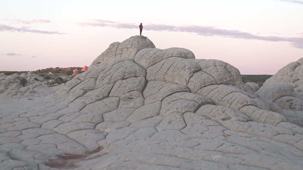 A drone flying forward and passing a male subject standing on top of the unique sandstone rock features of White Pocket Arizona surrounded by sandy desert and blue skies at blue hour sunrise