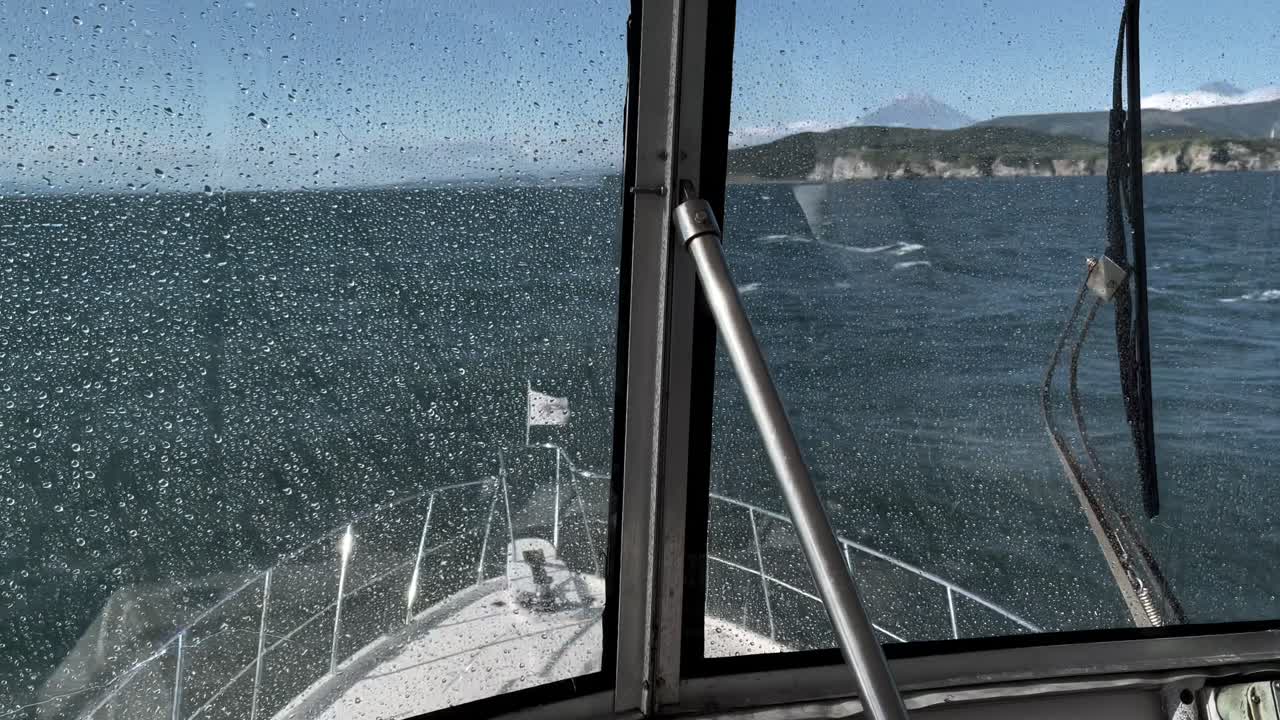 Blickwinkel aus dem Cockpit eines kleinen Bootes mit Regentropfen auf der Windschutzscheibe, über raue See fahrend auf eine zerklüftete Vulkan-Küste in Kamtschatka, Russland, an einem klaren Tag