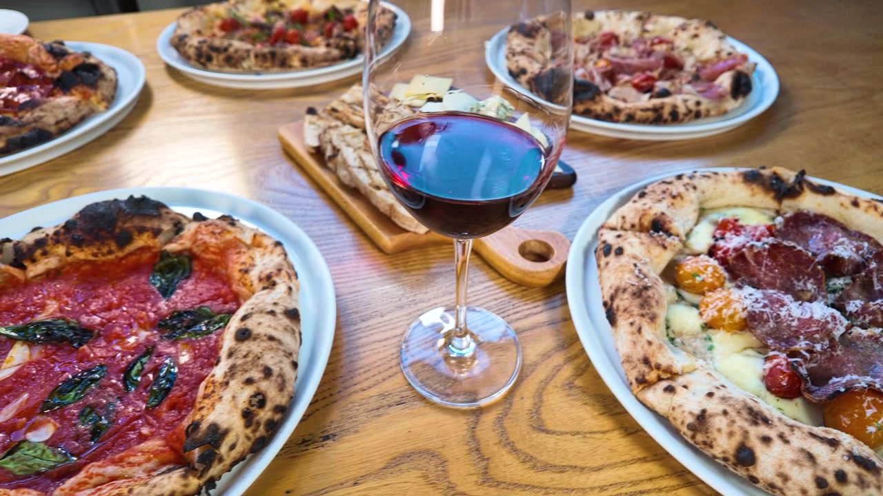 Ripresa panoramica dall'alto di pizze gourmet, vino rosso e antipasti su un tavolo di legno di un ristorante.