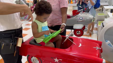 lovely two year old child in the hairdresser seated in a red toy car focused in an ipad next to his mother, wearing a sleeveless shirt