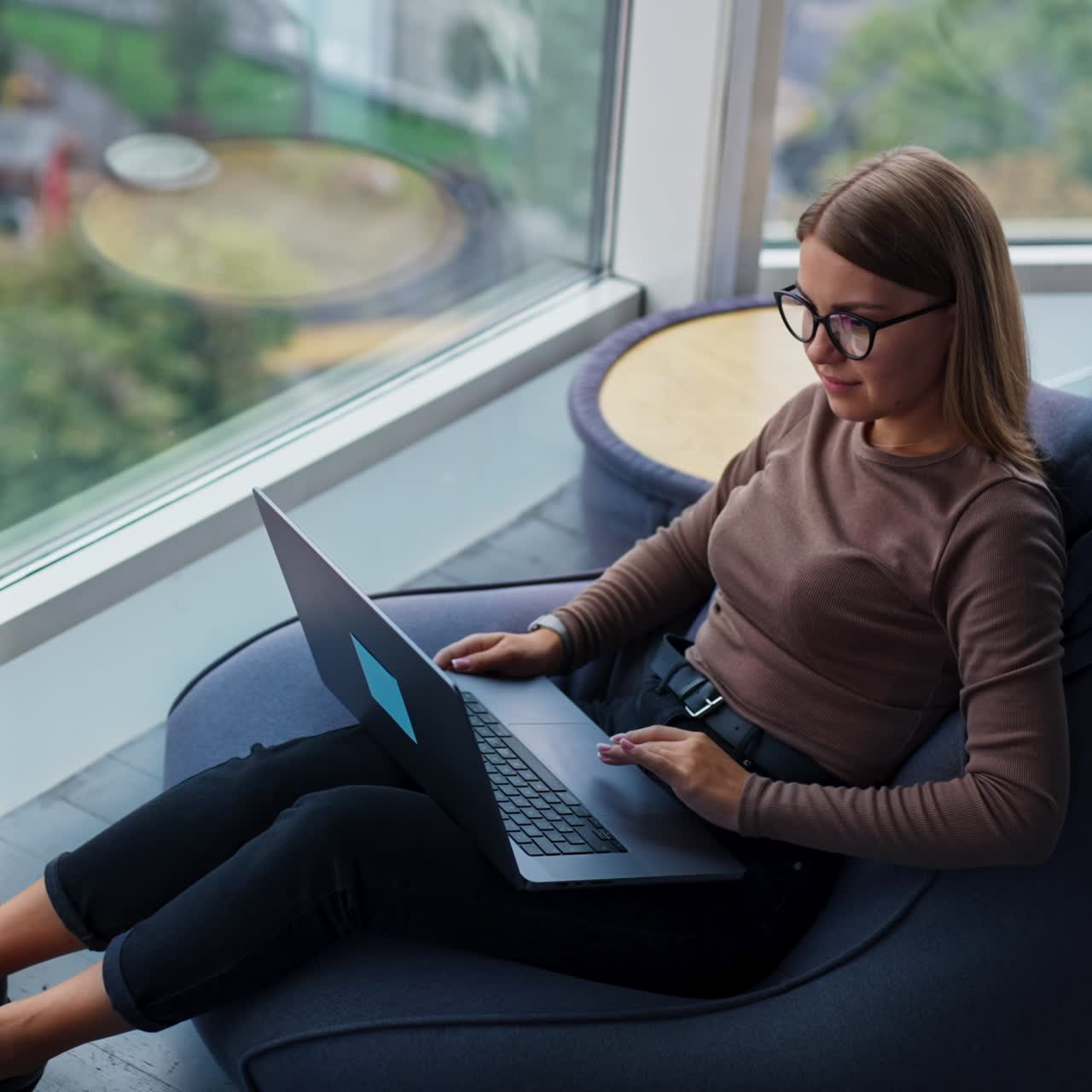 Thoughtful young lady sitting near big window opens her laptop and starts work. Woman typing on computer looking into window. High angle view