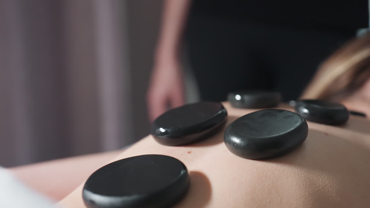 Very close up view of massage stones placed on client back during relaxing spa treatment with blurred massage therapist standing in background, highlighting warmth, amd calm atmosphere