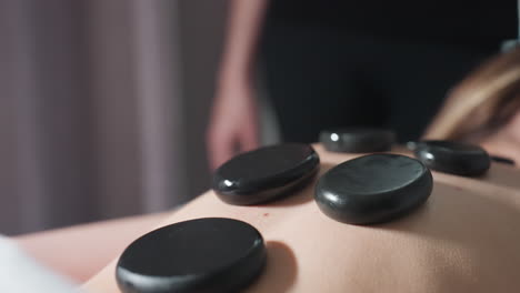 very close up view of massage stones placed on client back during relaxing spa treatment with blurred massage therapist standing in background, highlighting warmth, amd calm atmosphere