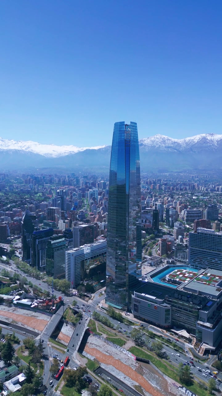Scatto panoramico di un imponente grattacielo di vetro che si erge sopra lo skyline della città con le Ande innevate sullo sfondo. Santiago, Cile