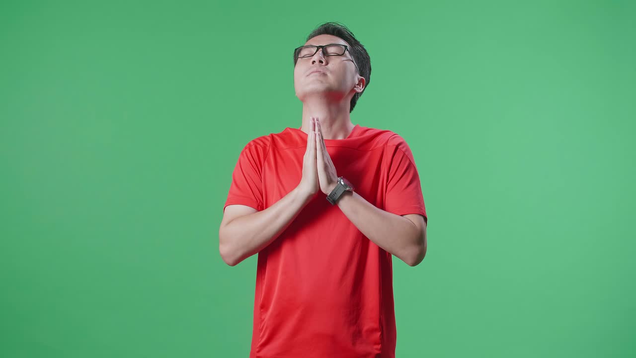 눈을 감고 공을 들고 있는 아시아인 남자가 초록색 스크린 배경에서 축구를 응원하는 동안 손을 들기 전에 팀을 위해 기도합니다.