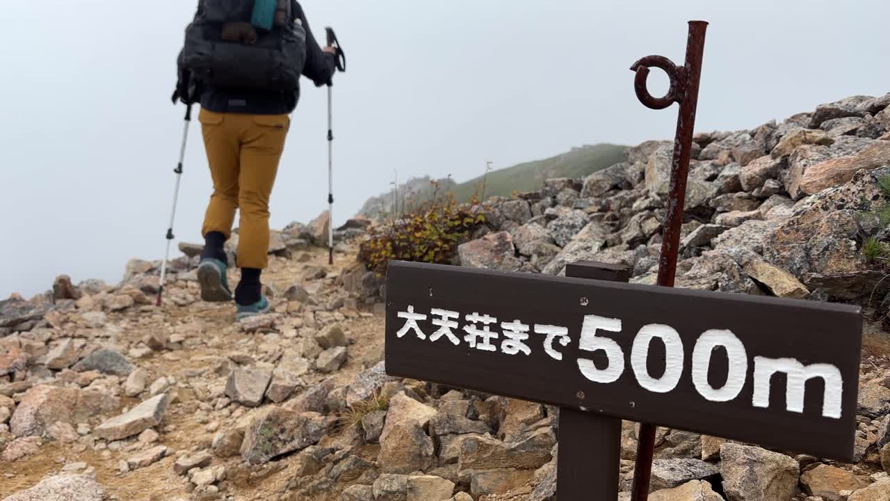 En kvindelig vandrer med rygsæk vandrer langs en stenet sti med en tåget baggrund i de Nordlige Japanske Alper, nær et skilt der angiver 500 meter til Daitenso Hytte.