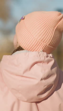 niña con gorra y chaqueta rosada, sosteniendo la mano de un adulto mientras camina en un parque iluminado por el sol, con árboles y edificios borrosos en el fondo