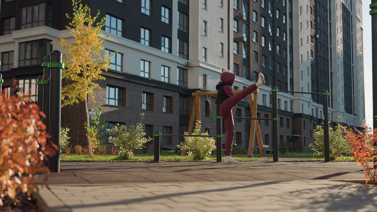 Fitness enthusiast holding pull up bar and stretching leg high beside metal bars in urban outdoor court under bright sun, wearing hoodie and sweatpants, modern residential background