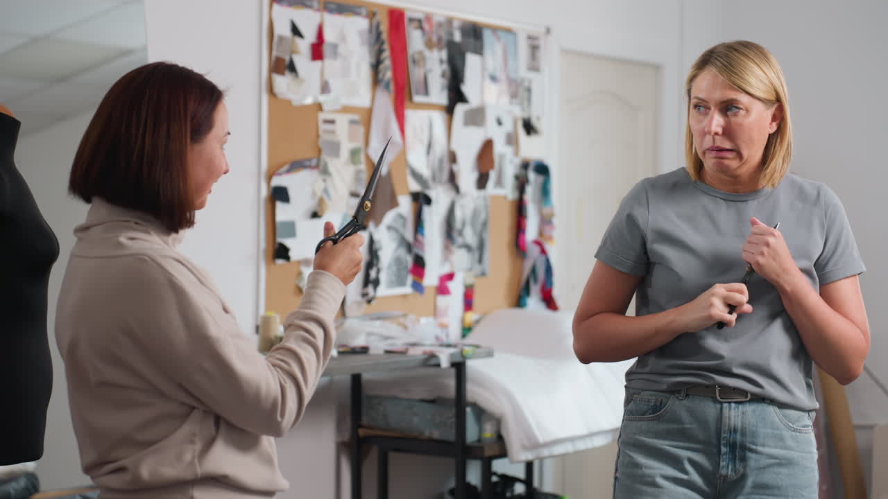 Seamstresses laughing and making playful sword fight gestures with large sewing scissors in lively creative workspace surrounded by colorful fabrics, design boards, sewing materials in background