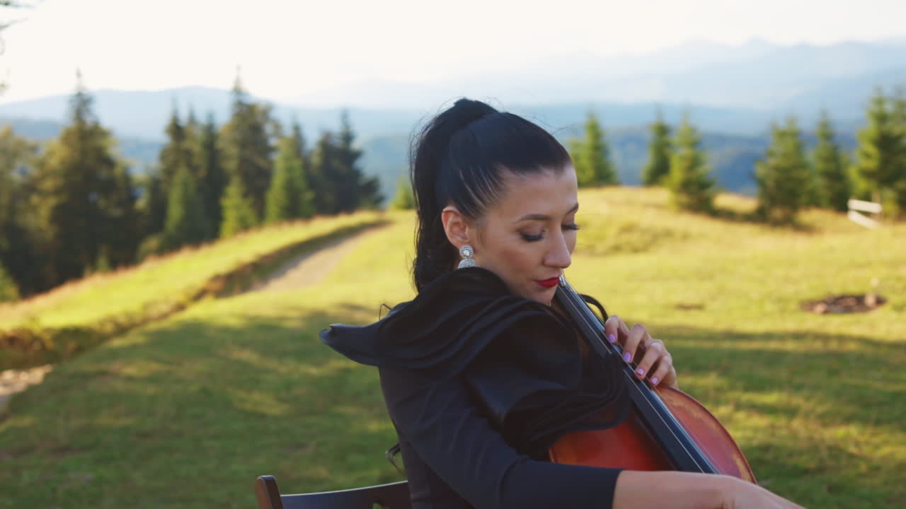 Musician with bright makeup performs classical music in the mountains. Woman playing cello sitting under the tree on the sunny meadow.
