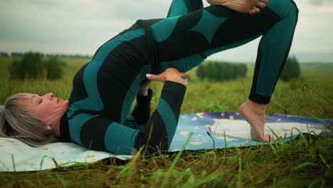 close-up of woman lying on yoga mat practicing bridge pose with hands supporting her waist, in a wide grassy field under cloudy skies, background shows blurred trees lined in the distance