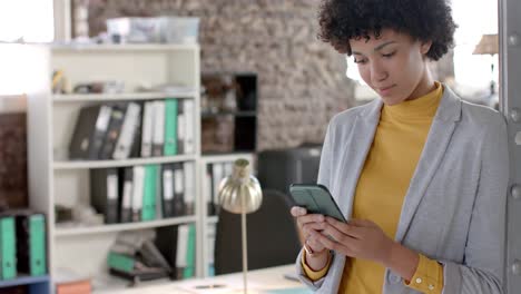 mujer de negocios casual biracial enfocada usando un teléfono inteligente en la oficina en cámara lenta