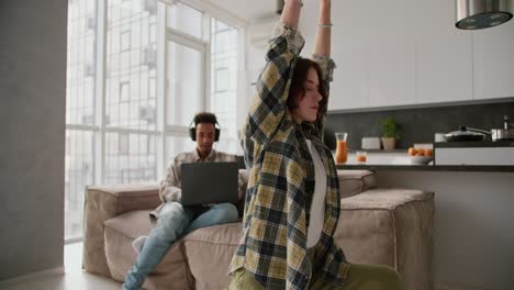 Una-Chica-De-Cabello-Castaño-Y-Peinado-Bob-Con-Una-Camisa-A-Cuadros-Verde-Hace-Yoga-Y-Deportes-Mientras-Su-Novio,-Un-Joven-De-Piel-Negra-Con-Auriculares-Negros,-Trabaja-En-Su-Computadora-Portátil-Gris-Sentado-En-El-Sofá-De-Un-Moderno-Apartamento-Tipo-Estudio.