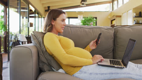 Happy-caucasian-pregnant-woman-sitting-in-armchair-and-using-smartphone-and-laptop