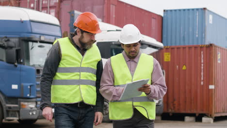 capo e lavoratore indossano giubbotti e caschi di sicurezza mentre organizzano una flotta di camion in un parco logistico mentre consultano un documento e camminano