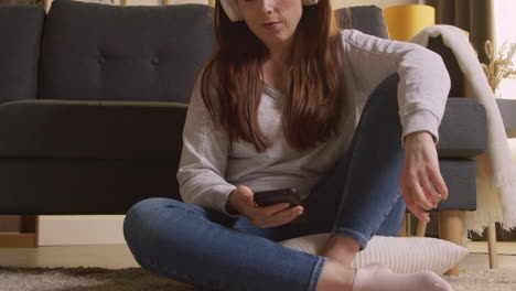 woman wearing wireless headphones sitting on floor at home streaming music from mobile phone 2