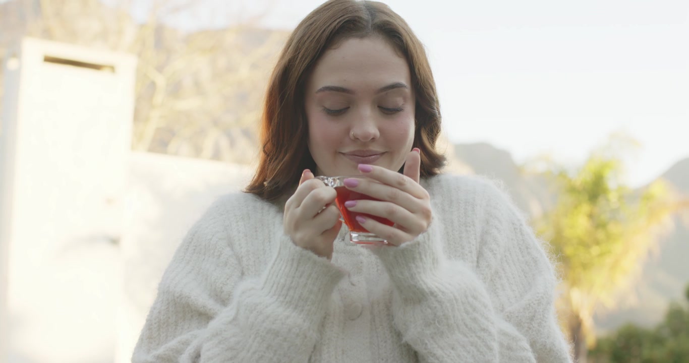 Donna adulta con cardigan spesso che porta la tazza da tè al viso, annusando il tè rosso e assaporandone l'aroma sul balcone.