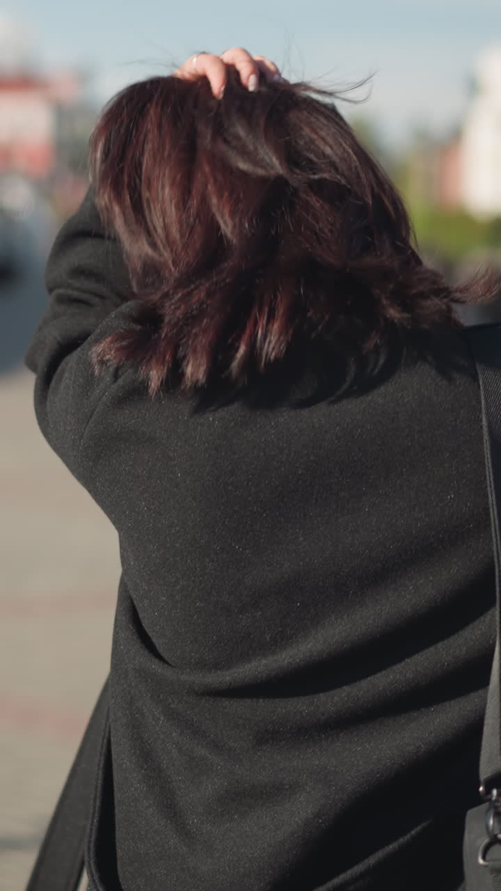 왼손으로 머리카락을 조정하고, 오른손에 검은 드백을 들고, 포장된 도시 거리를 고 있는 여성의 뒷면, 배경에는 흐릿한 건물, 초록색, 빨간색 경로가 있습니다.