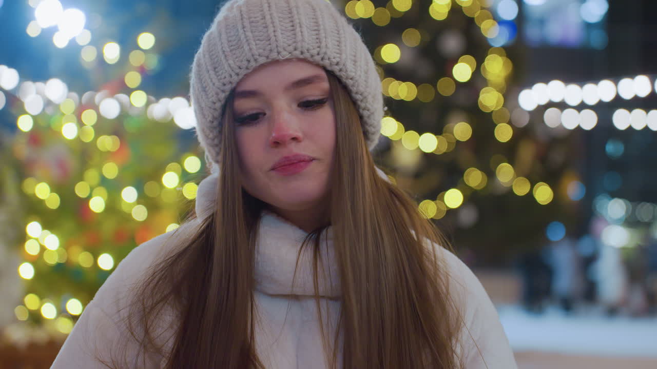 Close-up of woman in cozy winter coat and beanie walking toward a seat with head slightly bowed, festive city lights glow in background as people skate and socialize