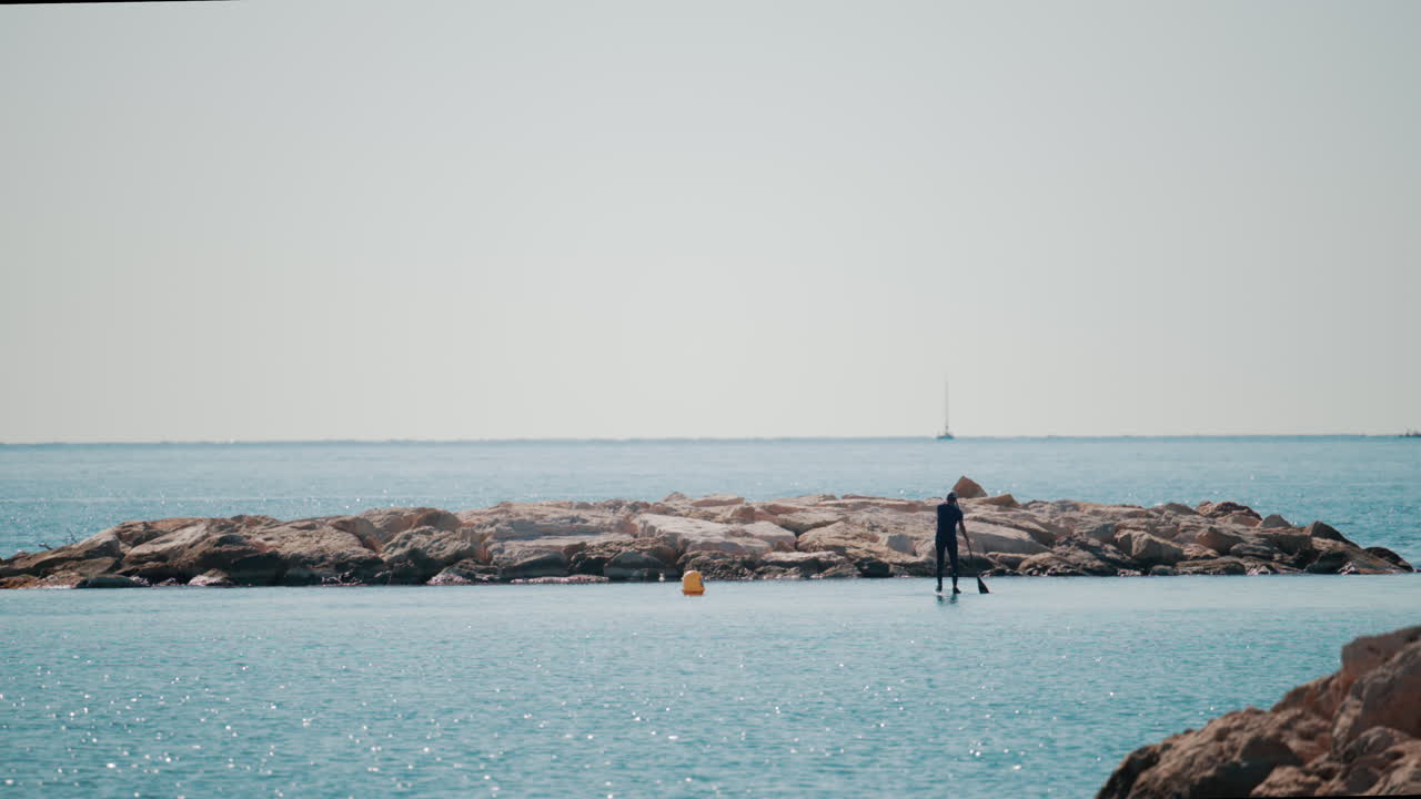 A man on a paddleboard approaches a rocky breakwater on a bright sunny day