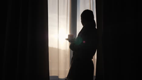 Die-Silhouette-Einer-Jungen-Frau,-Die-Morgenkaffee-Am-Fenster-Trinkt