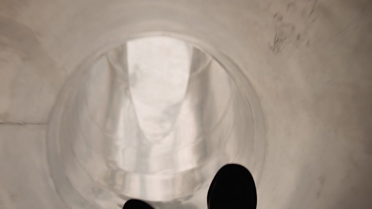 Pov feet on metal tube slide firstperson ride through curved stainless surface, sneakers and gray pants in frame, light reflections and shadowed tunnel, gradual descent building playful anticipation