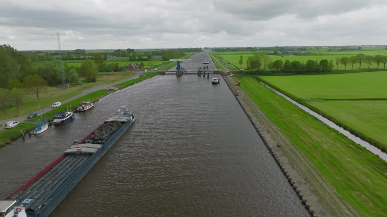 Wide reverse view past moored boats toward a bascule bridge spanning a straight canal through open farmland