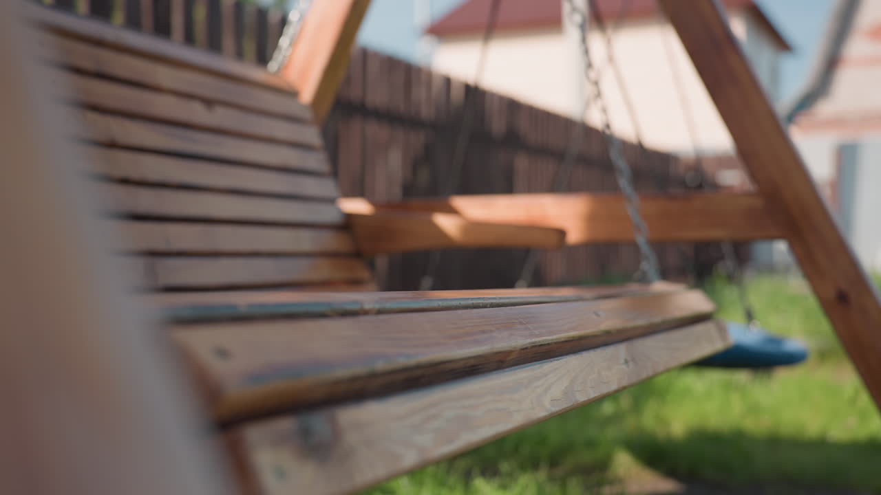 Close up of smooth wooden swing hanging by metal chain, gently swaying in bright backyard beside blue nest swing with soft grassy ground and blurred residential house in background
