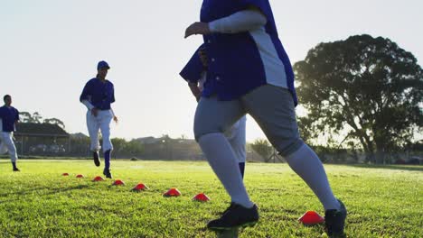 Diverse-group-of-female-baseball-players-exercising-on-sunny-pitch,-high-stepping-over-cones