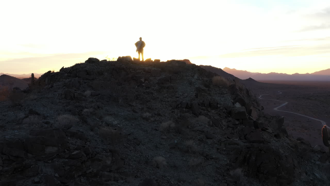 A dramatic silhouette of a lone man in a poncho, dressed as a spaghetti western hero, stands on a rugged mountain peak at sunset in the desert. The camera makes a pedestal up motion