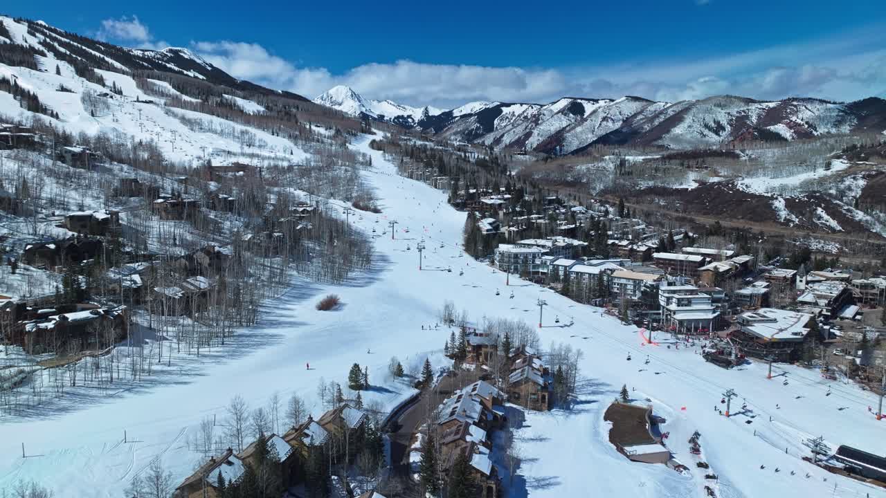 Ski trails wind down snowy forested slopes surrounded by mountain peaks and alpine terrain, skiers busy and lifts operate