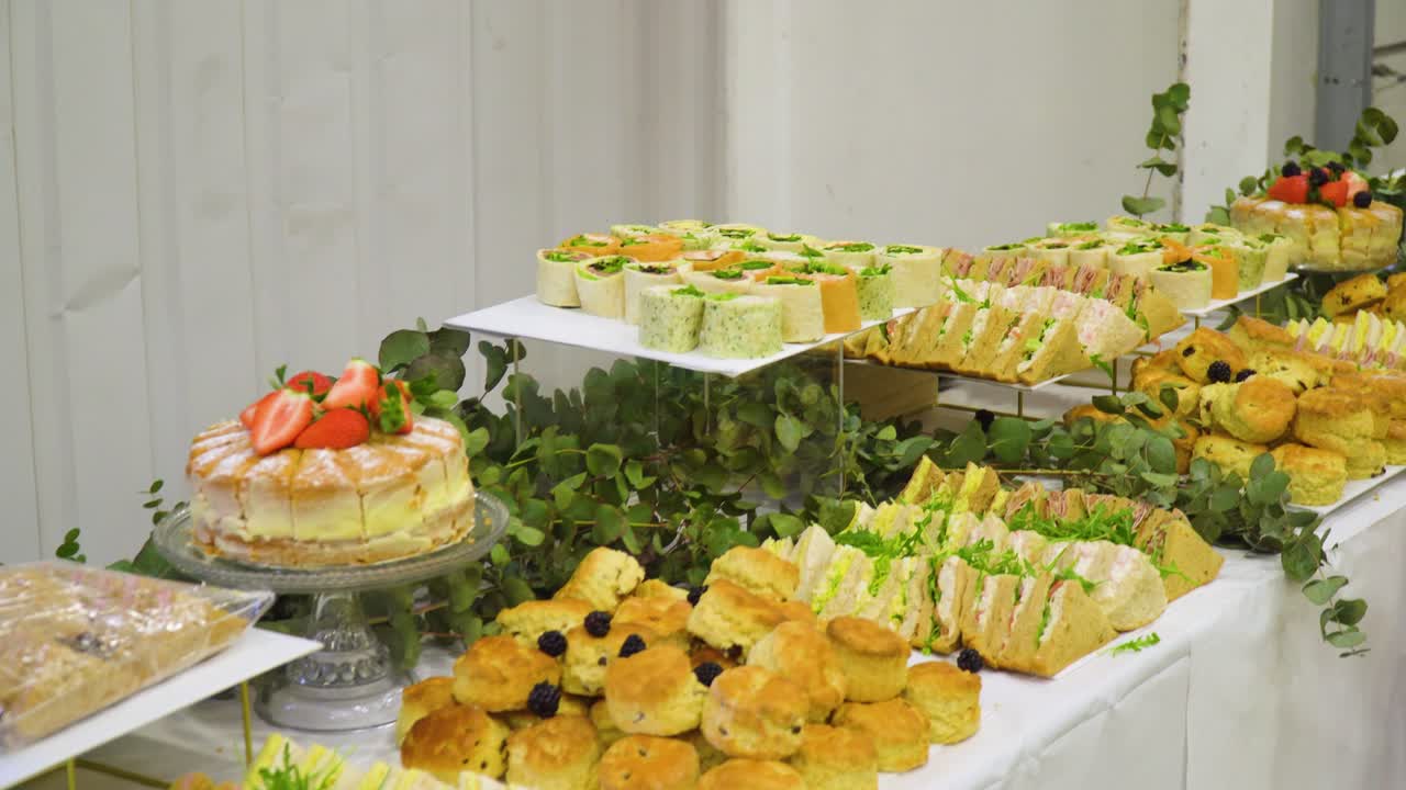 buffet inglés lleno de sándwiches y pasteles en una mesa larga dentro de un entorno de almacén para una reunión de negocios moviéndose hacia los lados revelando los alimentos y la selección ofrecida para el evento