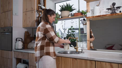 medium shot of woman filling jug with water from brass tap at kitchen sink surrounded by potted plants on window sill and reflected in wall mirror creating cozy greenery interior scene