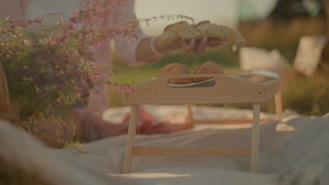 vista parcial de una mujer sentada en una alfombra de picnic al aire libre recogiendo croissants frescos de una bolsa de papel para colocarlos en platos encima de una mesa de picnic de madera, rodeada de una canasta tejida llena de flores