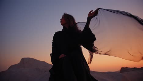 girl sitting on a rock in the desert wearing a black abaya and holding black transparent scarf wind blowing it during sunset in the fossil dunes