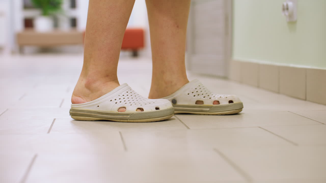 Rear leg view of health worker in white crocs walking confidently toward door in bright indoor corridor then shutting door behind her clean tiled hallway with visible reception couches