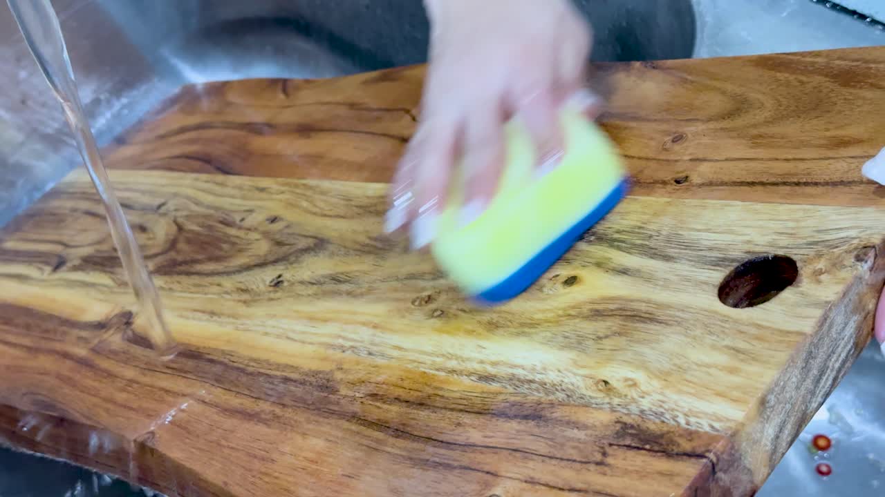 A person scrubs a large wooden cutting board with a sponge under a kitchen faucet. Bright lighting, close-up shot, steady camera, clean environment