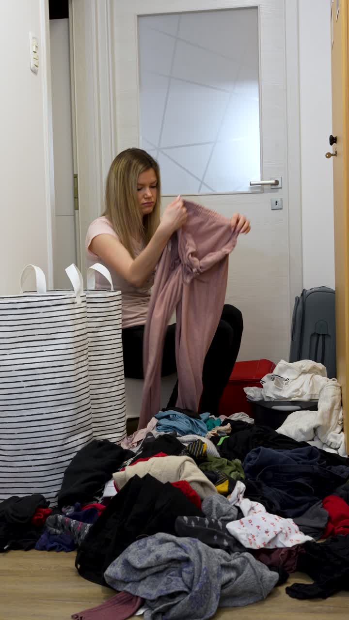 A young woman sits on the floor overwhelmed by a huge pile of messy clothes, sorting laundry into baskets after returning from a trip. Perfect for home life and domestic chore concepts