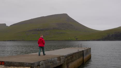 Woman-walking-towards-end-of-concrete-platform-in-Bour,-Faroe-landscape,-wide